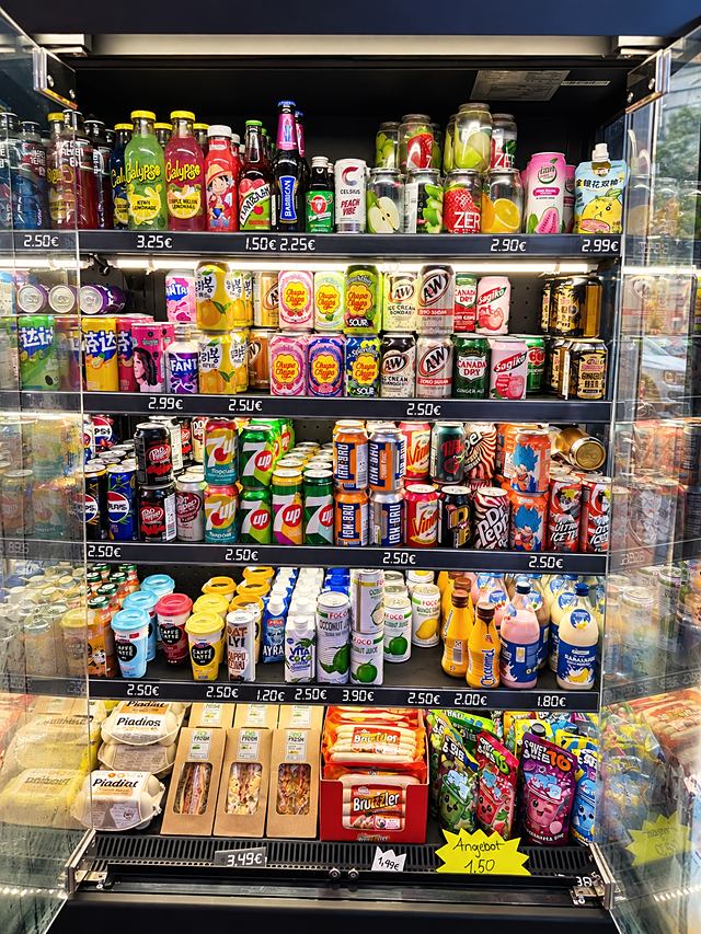 Offener Getränkekühlschrank mit bunten Dosen und Flaschen, Preisschildern und einem handgeschriebenen Angebot für 1,50 Euro.